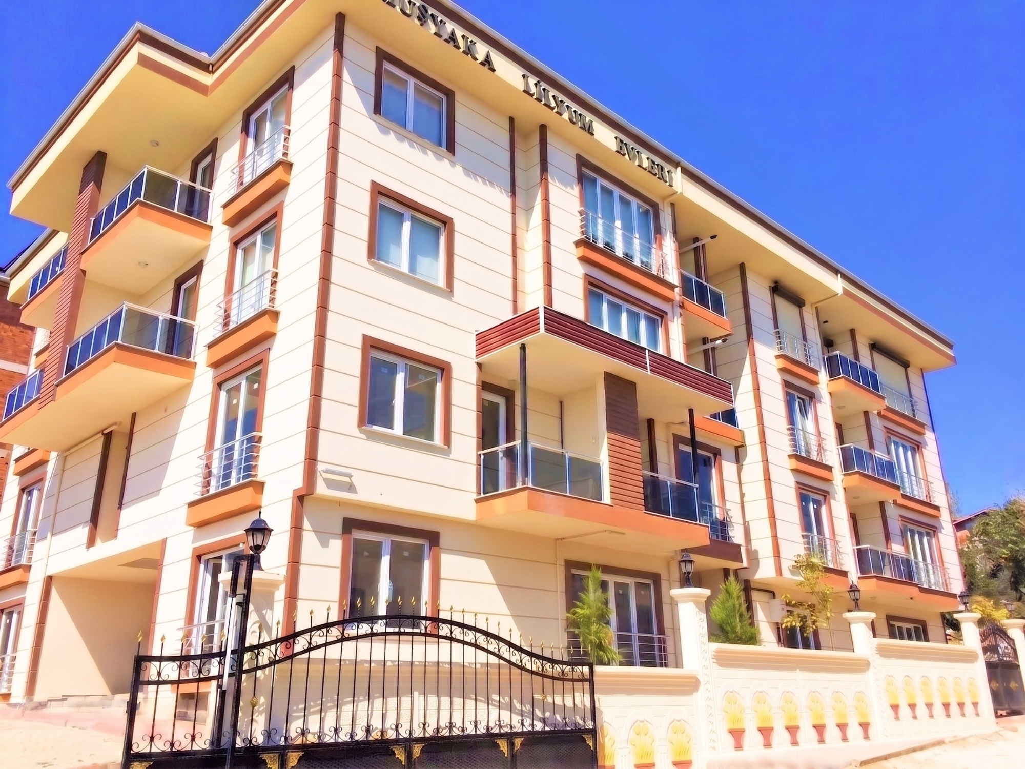 Dört katlı apartmanın bej-kırmızı cephesi ve ferforje bahçe kapısı — Gümüşyaka Konut Projesi I